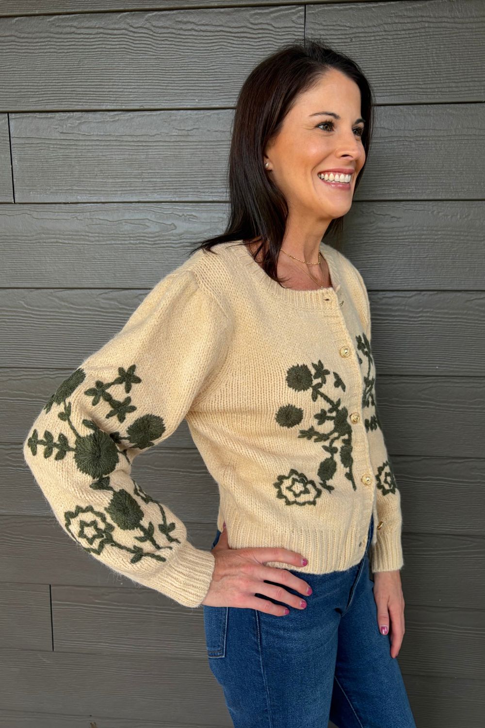 A woman wearing a cream-colored knit cardigan with dark green floral and cross motifs on the sleeves and body, paired with blue jeans, standing against a gray wood-paneled wall.
