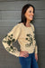 A woman wearing a cream-colored knit cardigan with dark green floral and cross motifs on the sleeves and body, paired with blue jeans, standing against a gray wood-paneled wall.