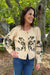 A woman wearing a cream-colored knit cardigan with dark green floral patterns and blue jeans, standing outdoors in front of a tree.
