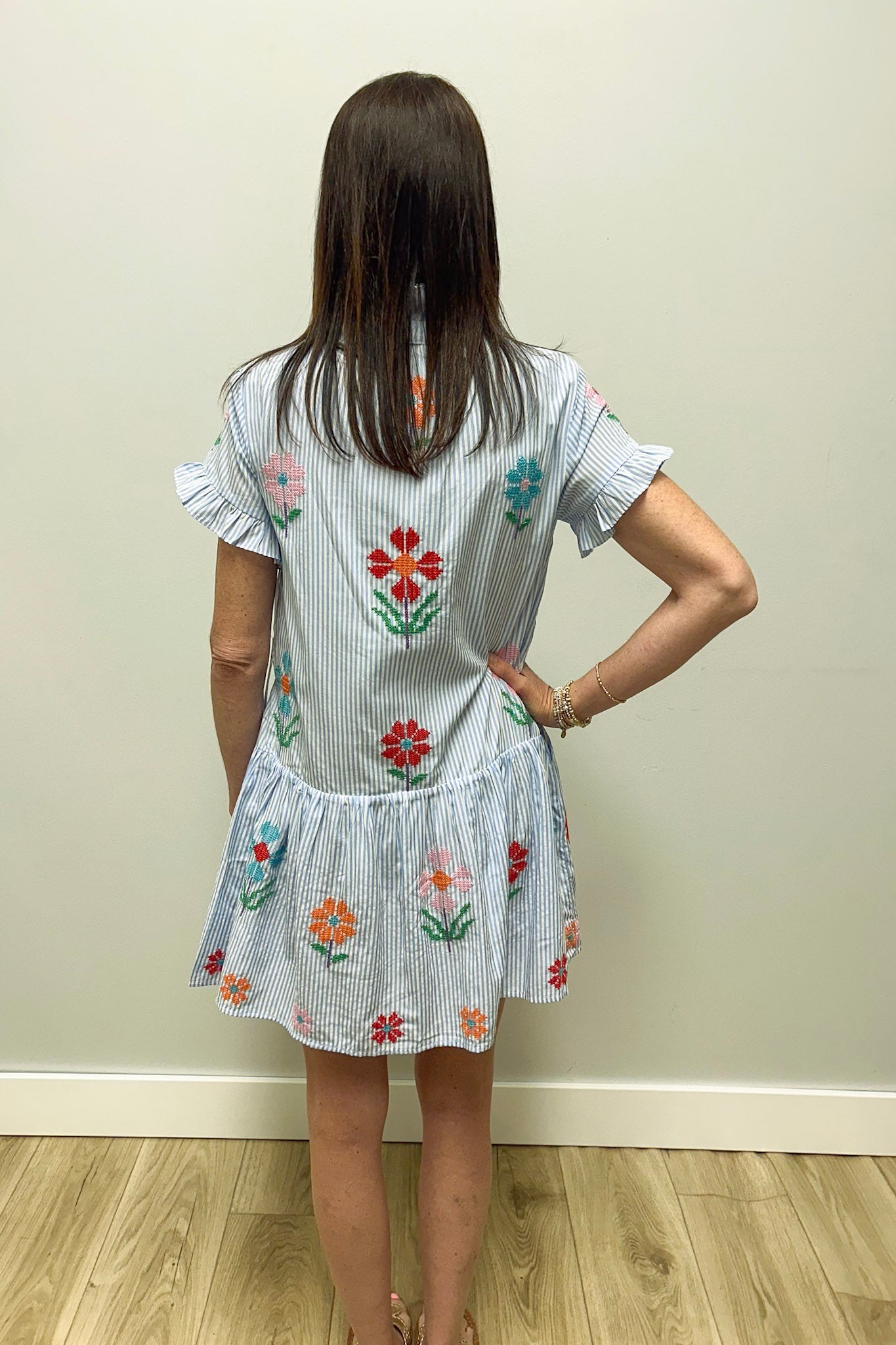 Woman wearing a white stripe dress with floral embroidery against a plain wall.
