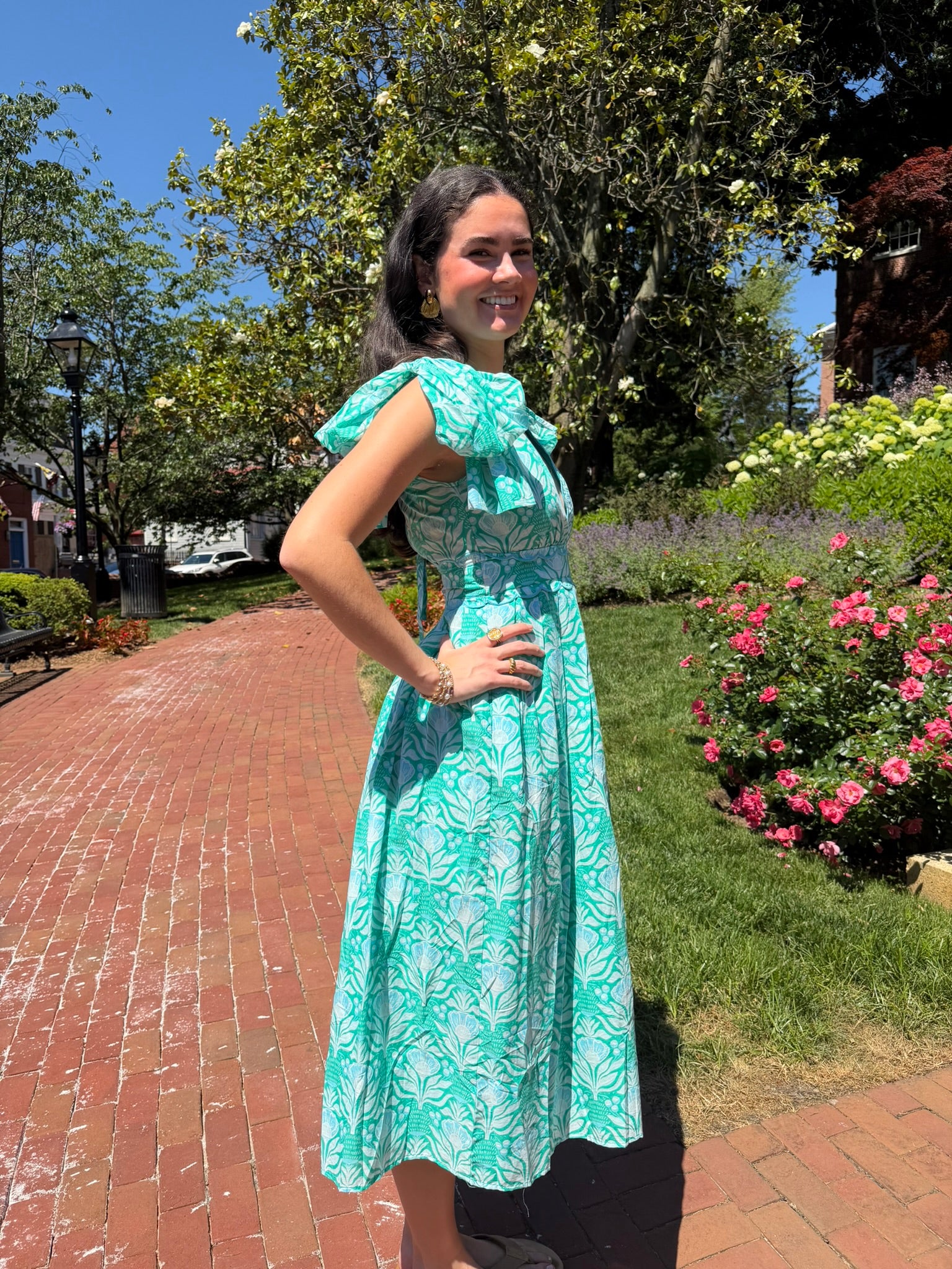 Woman in a blue dress standing outdoors on a brick path with trees and flowers in the background