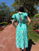 Woman in a green dress standing on a brick path with trees and flowers in the background
