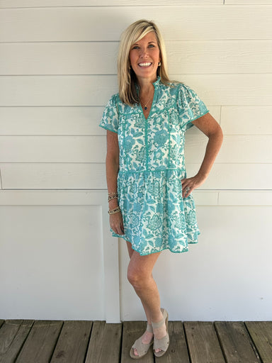 Woman wearing a turquoise floral dress standing against a white wooden wall.
