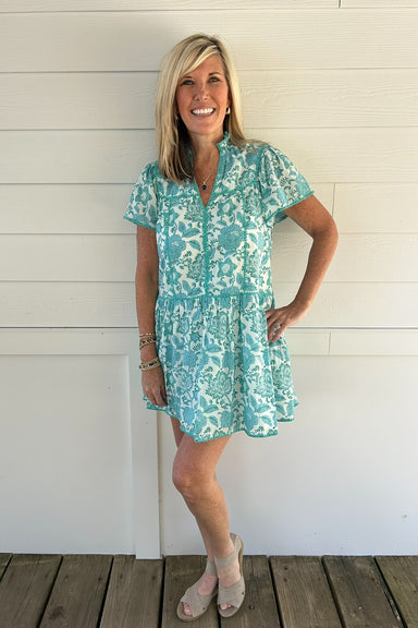 Woman wearing a turquoise floral dress standing against a white wooden wall.