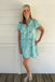 Woman wearing a turquoise floral dress standing against a white wooden wall.