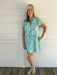 Woman wearing a turquoise floral dress standing against a white wooden wall.