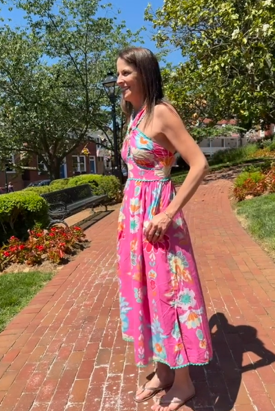 Woman in a pink floral dress standing on a brick path with trees and buildings in the background