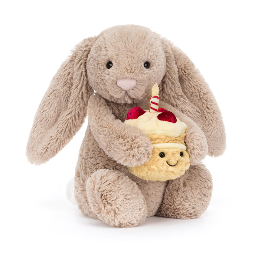 Jellycat Bashful Beige Bunny 'Birthday' is a beige plush bunny holding a plush double layer yellow cake topped with red strawberries, cream frosting, and a lit red and white striped candle. Front facing sitting