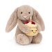 Jellycat Bashful Beige Bunny 'Birthday' is a beige plush bunny holding a plush double layer yellow cake topped with red strawberries, cream frosting, and a lit red and white striped candle. Front facing sitting