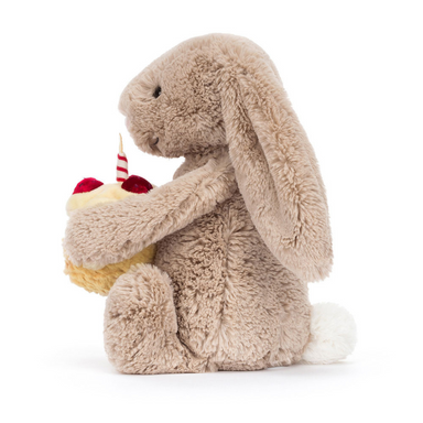 Jellycat Bashful Beige Bunny 'Birthday' is a beige plush bunny holding a plush double layer yellow cake topped with red strawberries, cream frosting, and a lit red and white striped candle. Side facing sitting