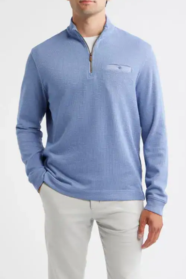 A man wearing a light blue quarter-zip knit sweater with a chest pocket, paired with white pants, against a plain light gray background.