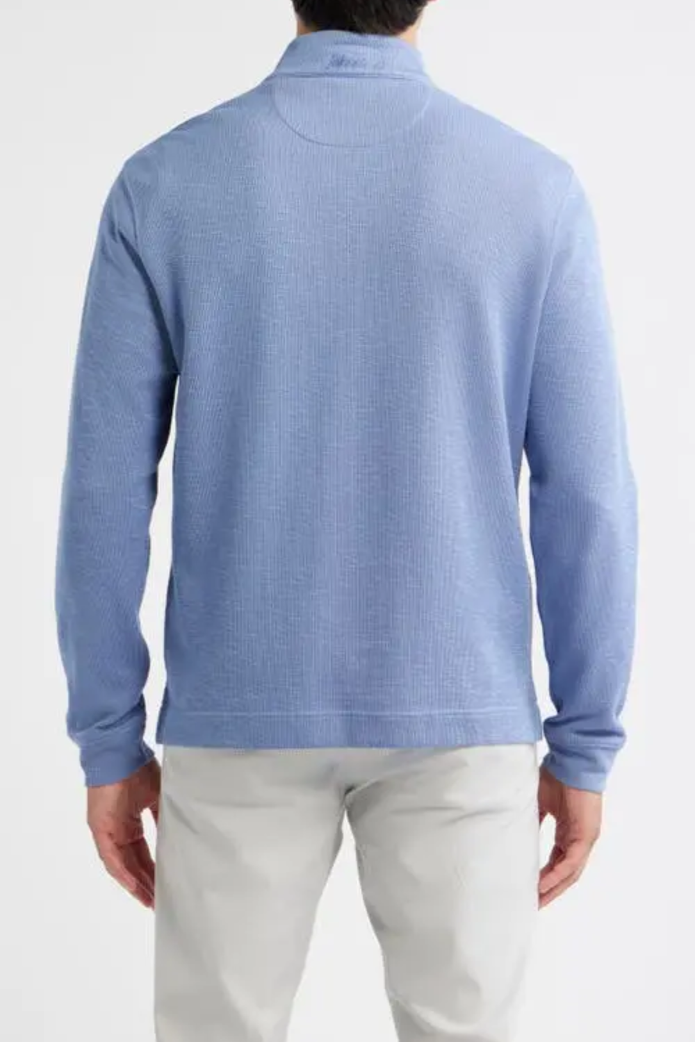 A man viewed from the back wearing a light blue ribbed knit quarter-zip sweater and off-white trousers, against a plain white studio background.