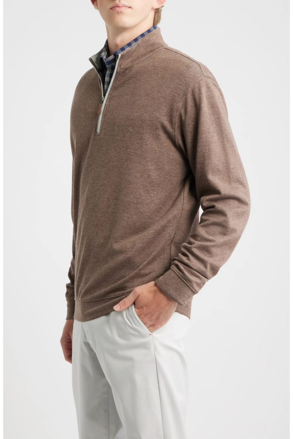 A man wearing a brown long-sleeve quarter-zip pullover sweater over a patterned collared shirt and light gray trousers, photographed against a plain light gray studio background.