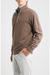 A man wearing a brown long-sleeve quarter-zip pullover sweater over a patterned collared shirt and light gray trousers, photographed against a plain light gray studio background.