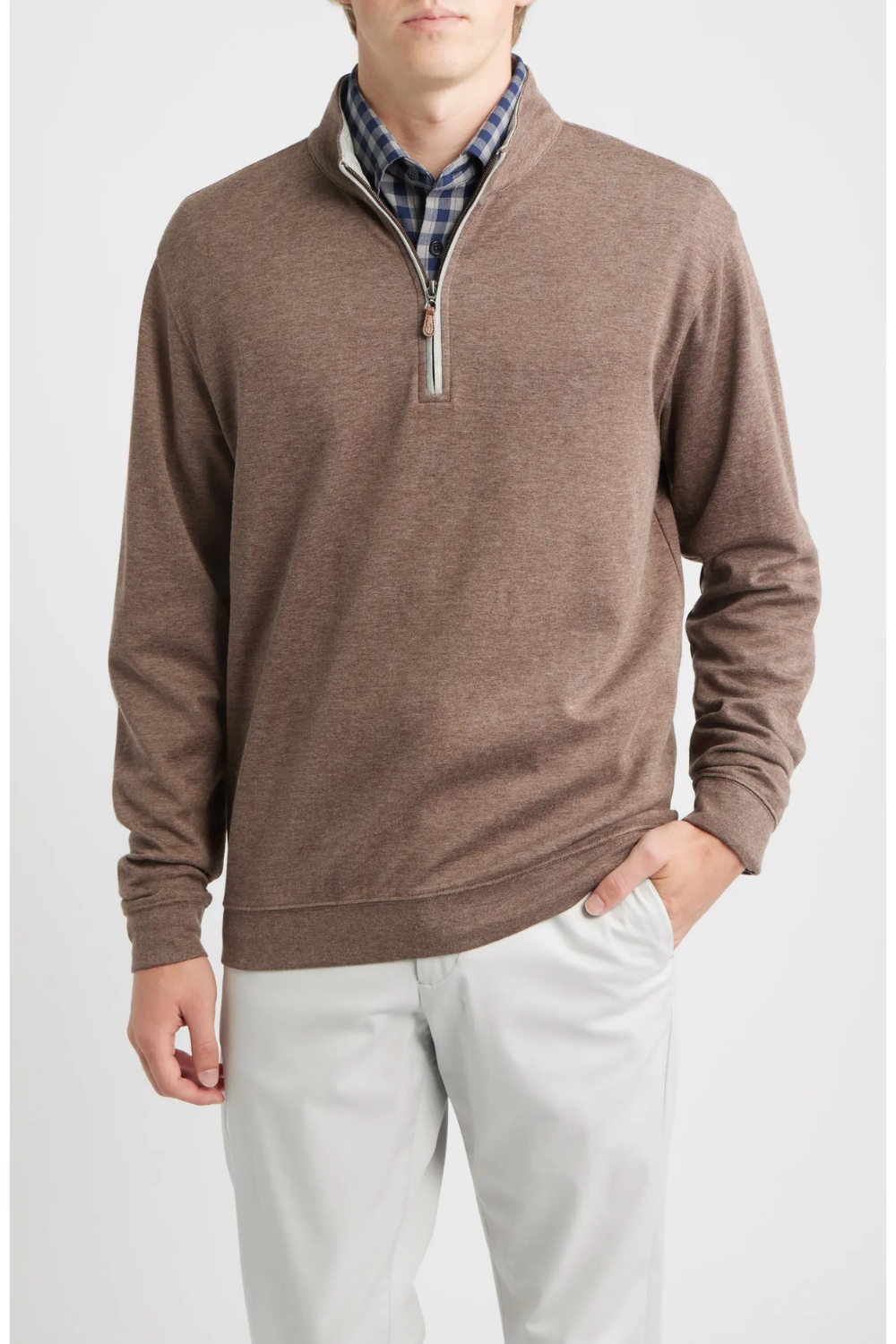 A man wearing a brown heathered quarter-zip pullover sweater layered over a blue and white plaid shirt, paired with light gray trousers, against a light gray studio background.