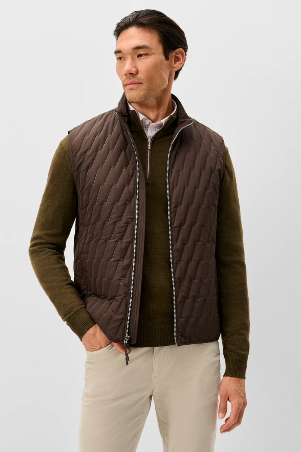 A man wearing a dark brown quilted zip-front vest over an olive green sweater and light beige pants, photographed against a plain light gray studio background.
