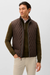 A man wearing a dark brown quilted zip-front vest over an olive green sweater and light beige pants, photographed against a plain light gray studio background.