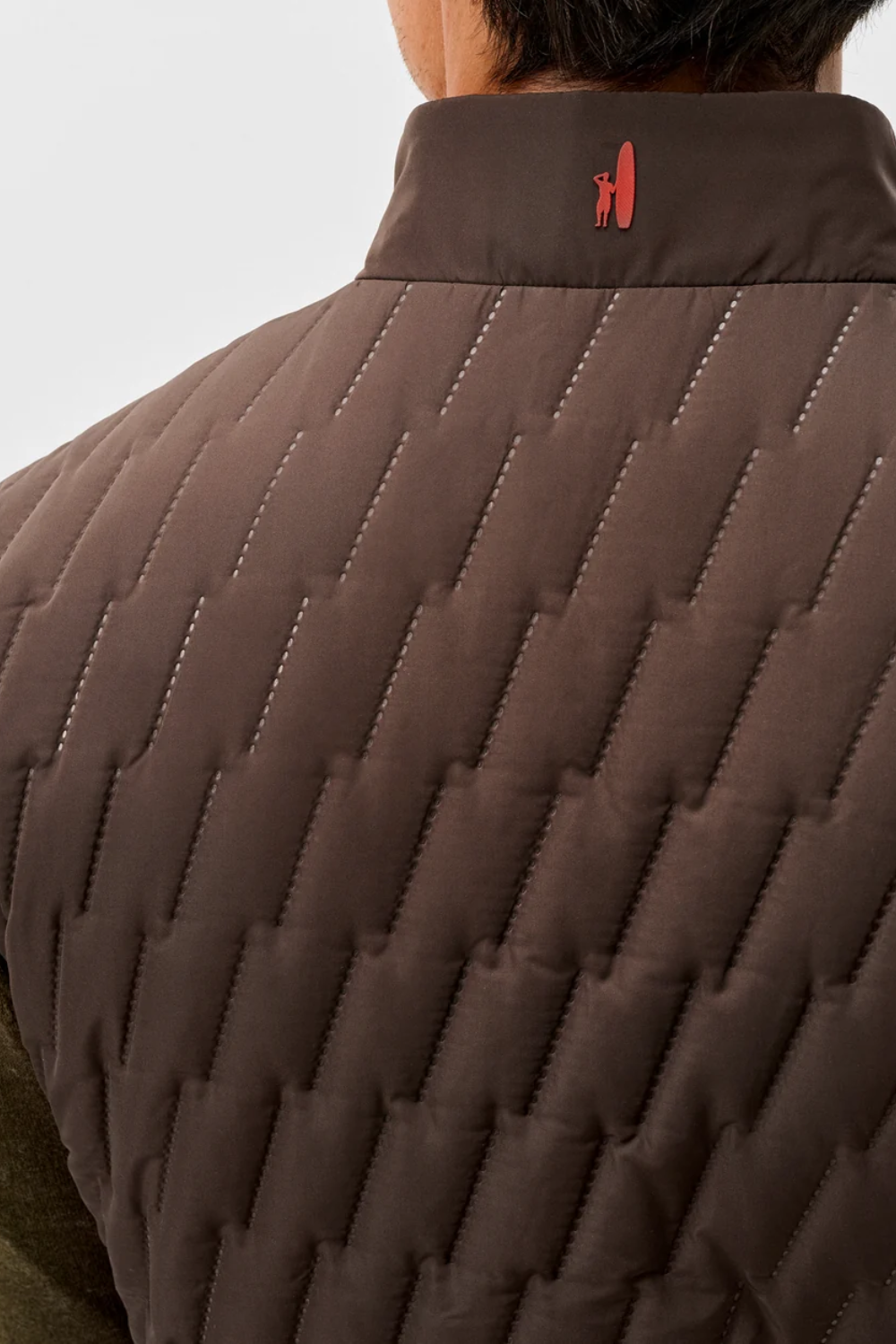 Close-up back view of a person wearing a brown quilted puffer vest with a high collar and a small red embroidered logo on the neck.