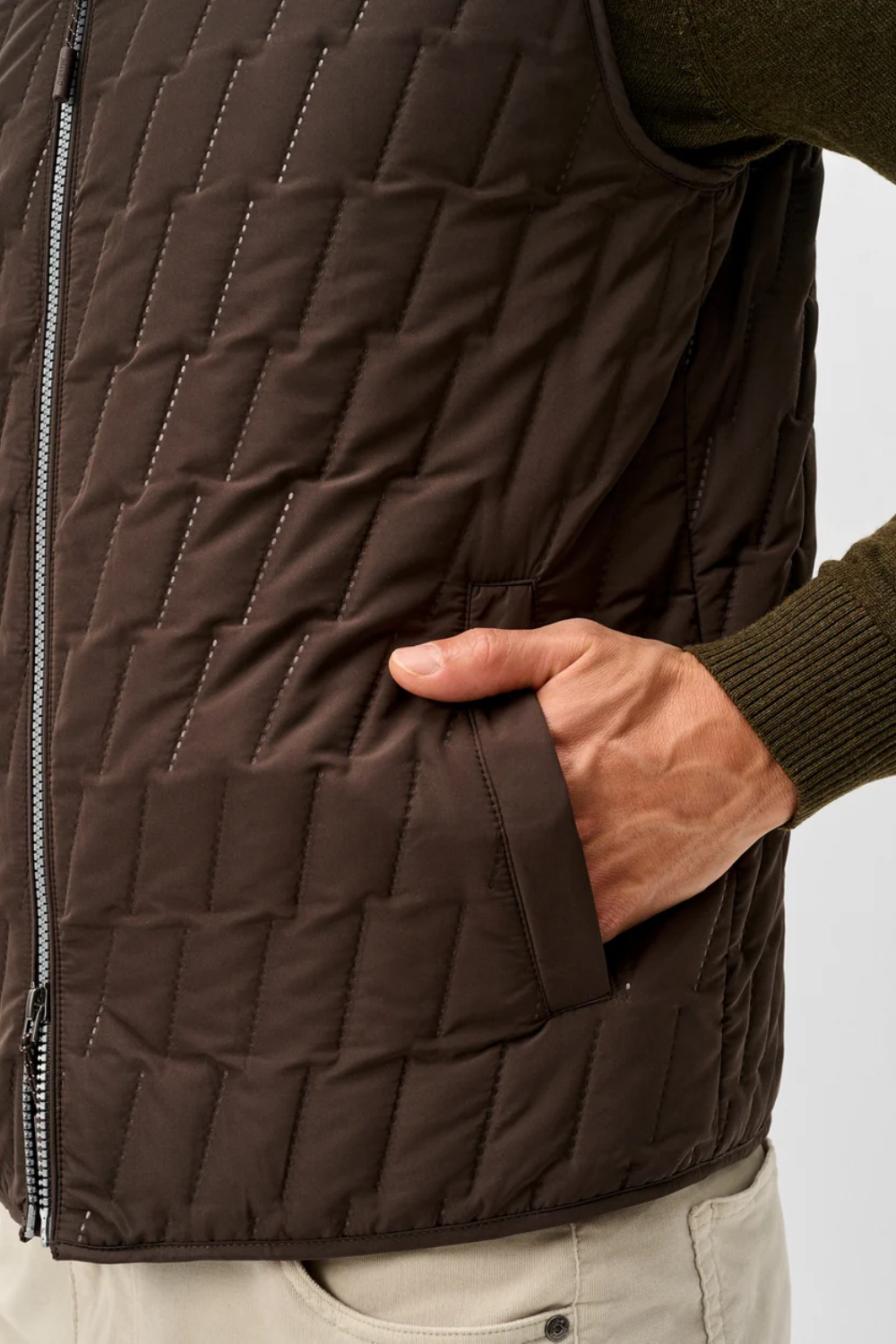 Close-up of a dark brown quilted puffer vest worn by a model, showing a hand in the side pocket and a zippered front closure.