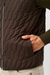 Close-up of a dark brown quilted puffer vest worn by a model, showing a hand in the side pocket and a zippered front closure.