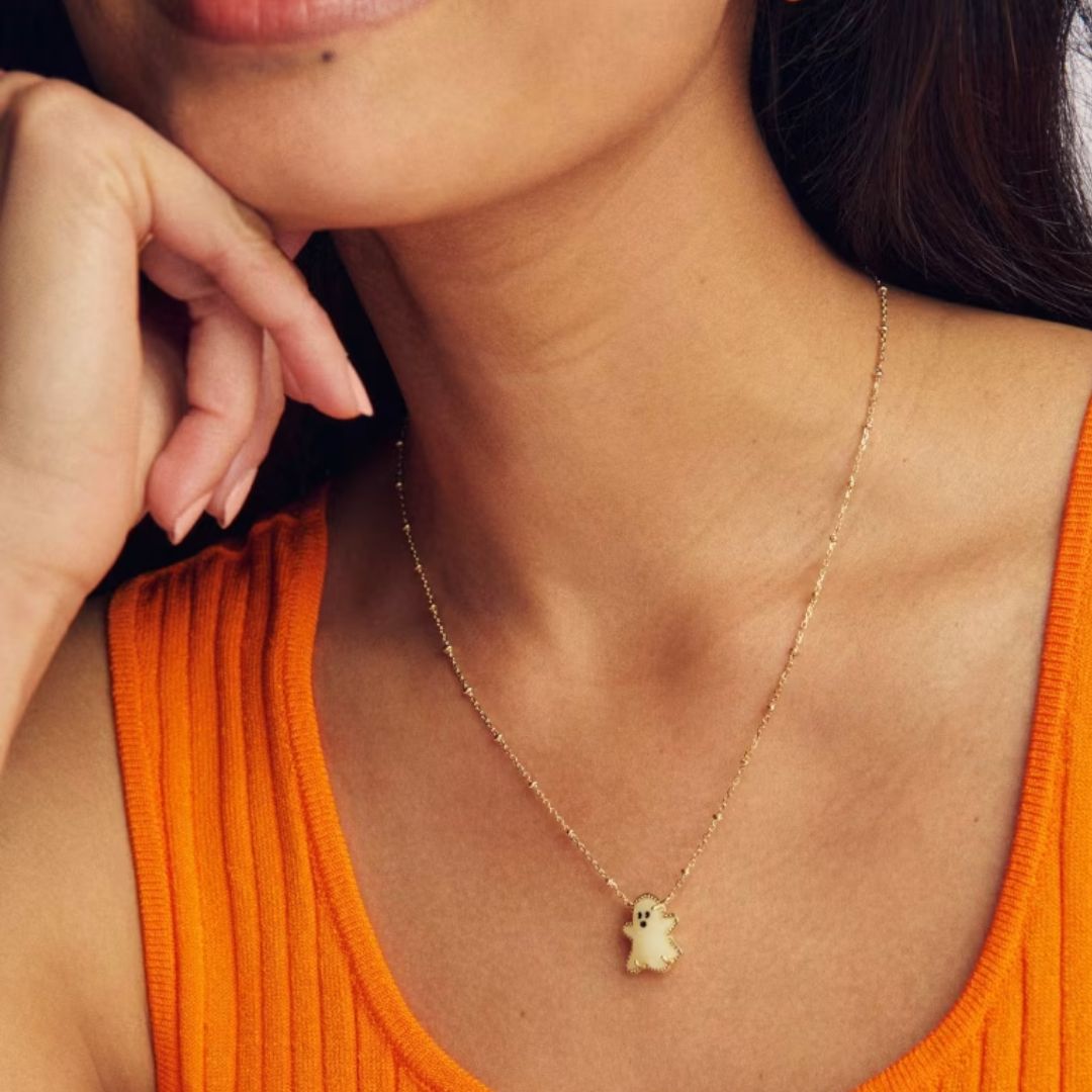 A close-up lifestyle shot of a model wearing a delicate gold chain necklace with a small cream-colored ghost pendant, styled with an orange ribbed tank top.