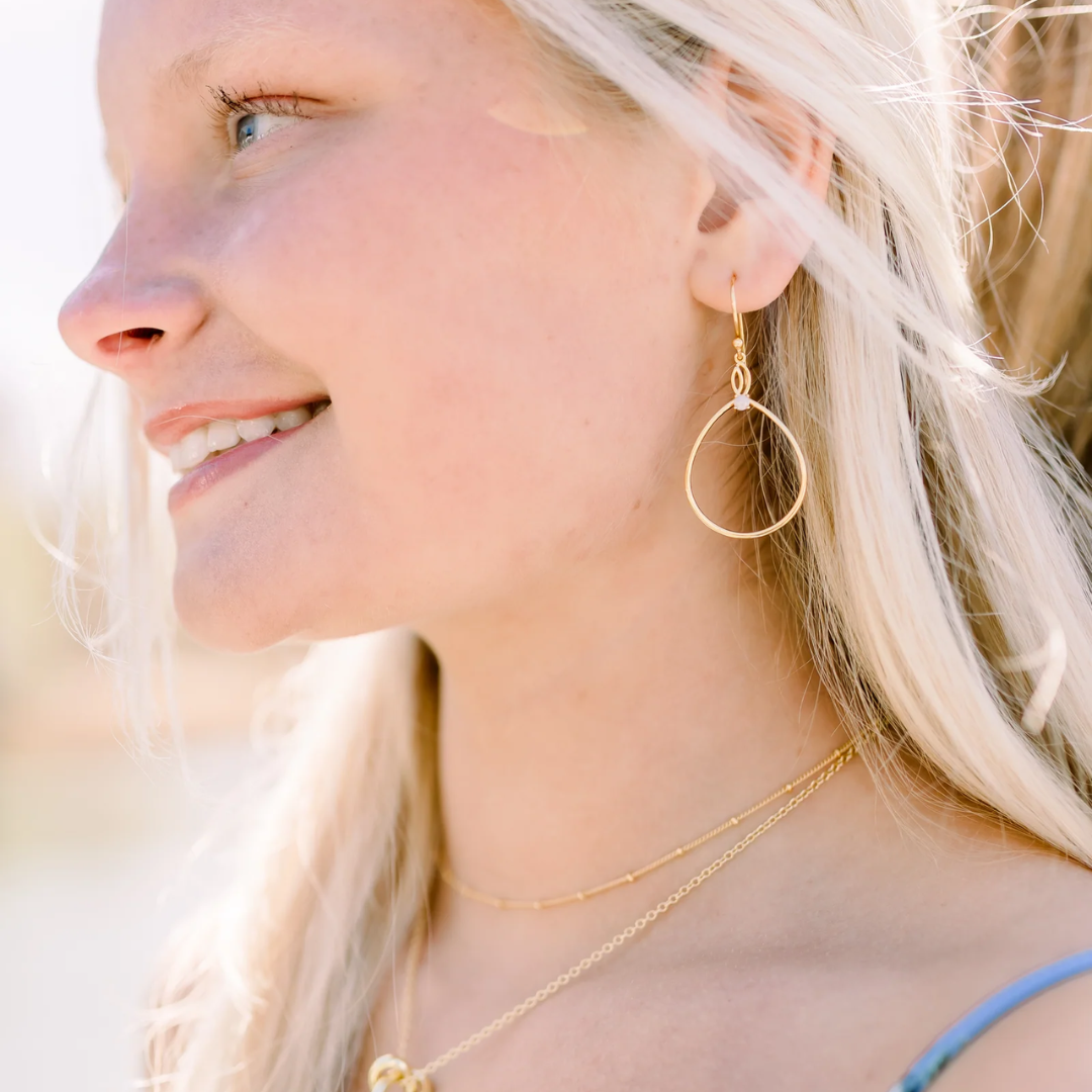 A close-up side profile of a smiling blonde woman wearing a delicate gold hoop earring and layered gold necklaces.