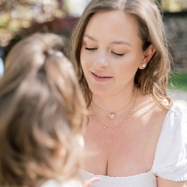 A woman wearing delicate gold necklaces, including a small heart-shaped pendant, outdoors with a child in the foreground.