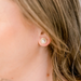 A close-up of a person's ear wearing a small gold hoop earring with a white pearl at the center, with light brown hair visible.