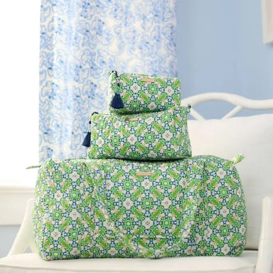 Three quilted travel or toiletry bags stacked on a white chair, featuring a green, white, and blue geometric floral pattern with dark blue tassels on the zippers.