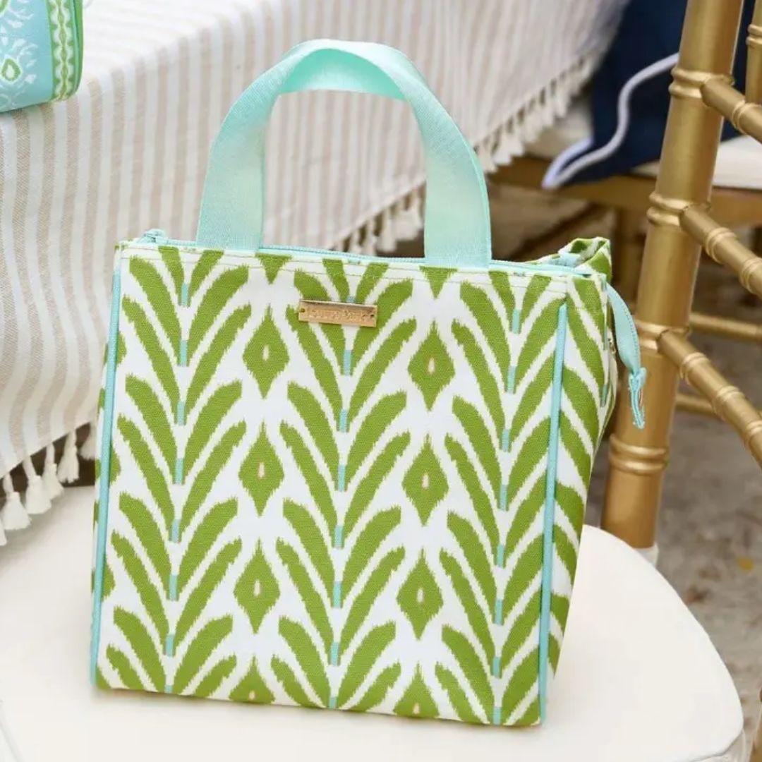 A square canvas lunch bag with a green and white ikat-style leaf pattern, light aqua handles and piping, and a small gold-tone brand plaque on the front, shown resting on a white chair in a styled indoor setting.