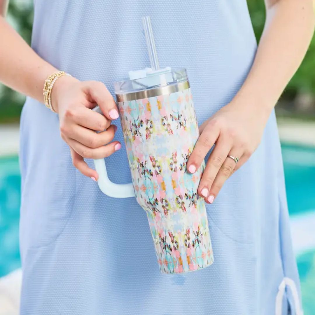 A person in a light blue dress holds a handled tumbler with a pastel multicolor pattern and a clear straw, with a swimming pool and greenery blurred in the background.