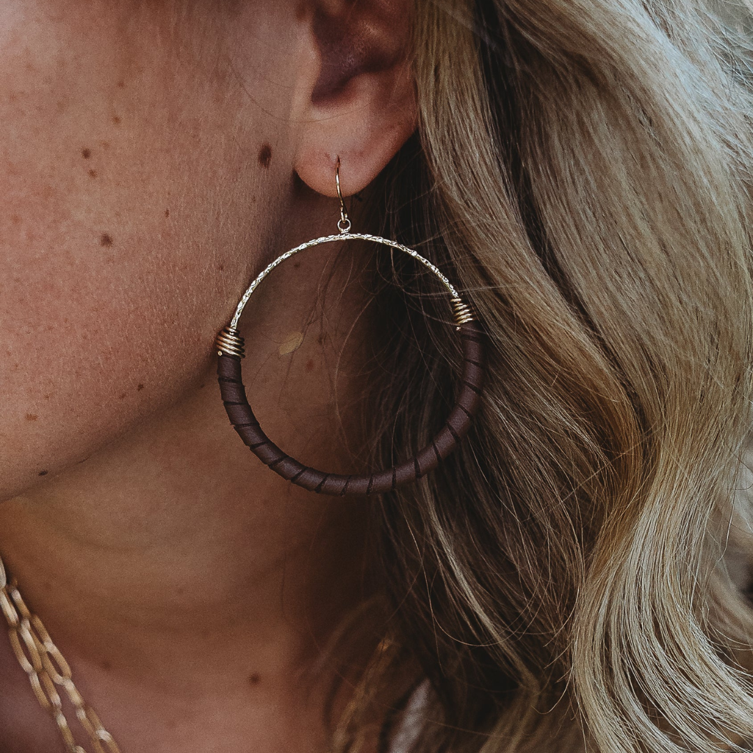 A close-up of a person wearing a large hoop earring. The earring has a dark brown textured wrap with gold-toned metal wire accents and a gold-toned hook.