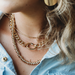 A close-up of a person wearing layered gold necklaces, including a chunky curb chain, a delicate thin chain, and a beaded chain, paired with a light blue denim shirt.
