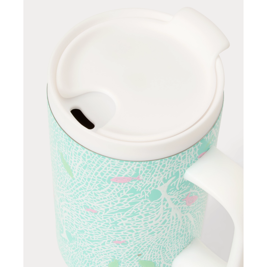 A close-up of a ceramic mug with a white handle and lid, featuring a mint green and white tree pattern with small pink accents.