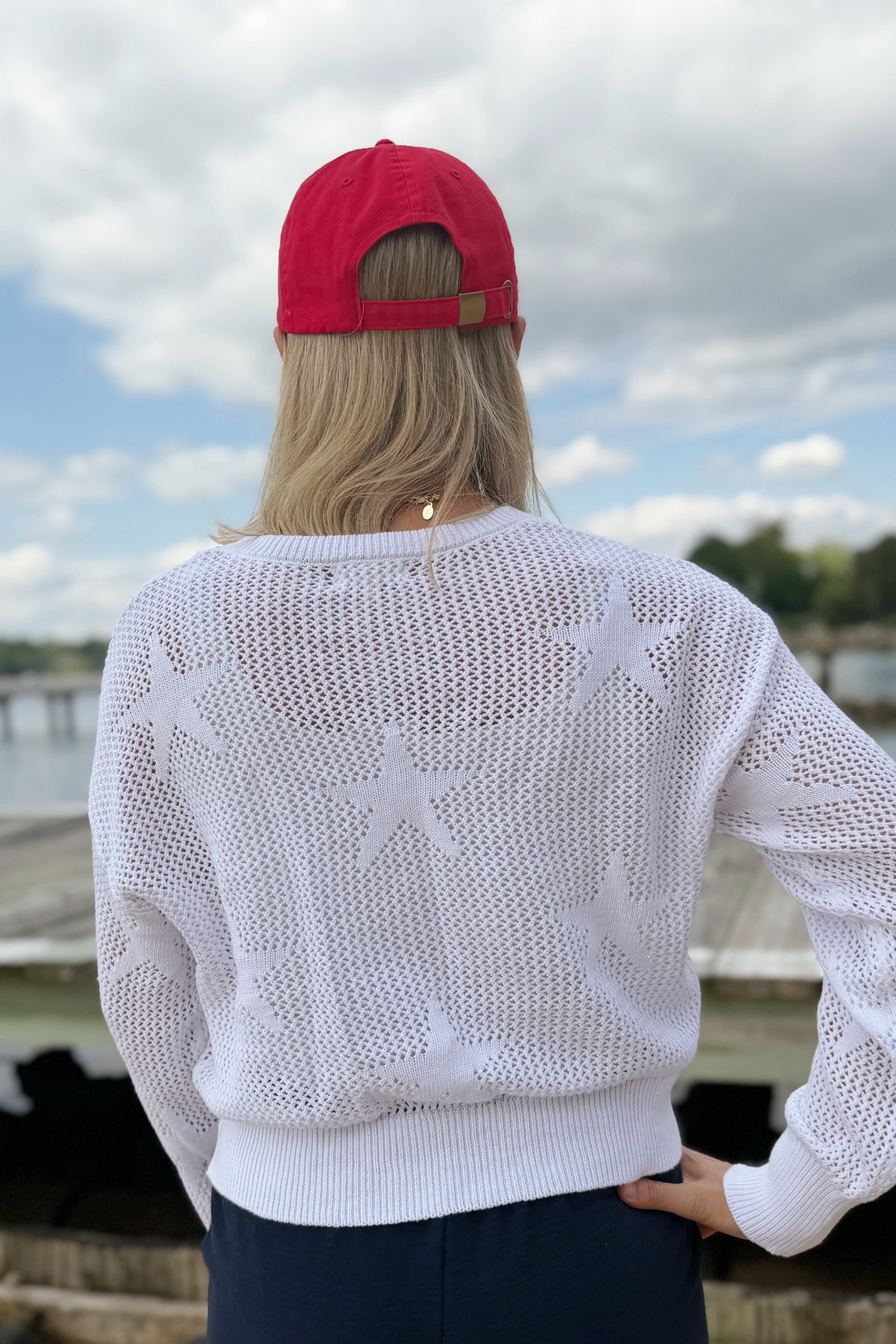 Person wearing a white sweater with star patterns and a red cap, standing outdoors by a body of water.