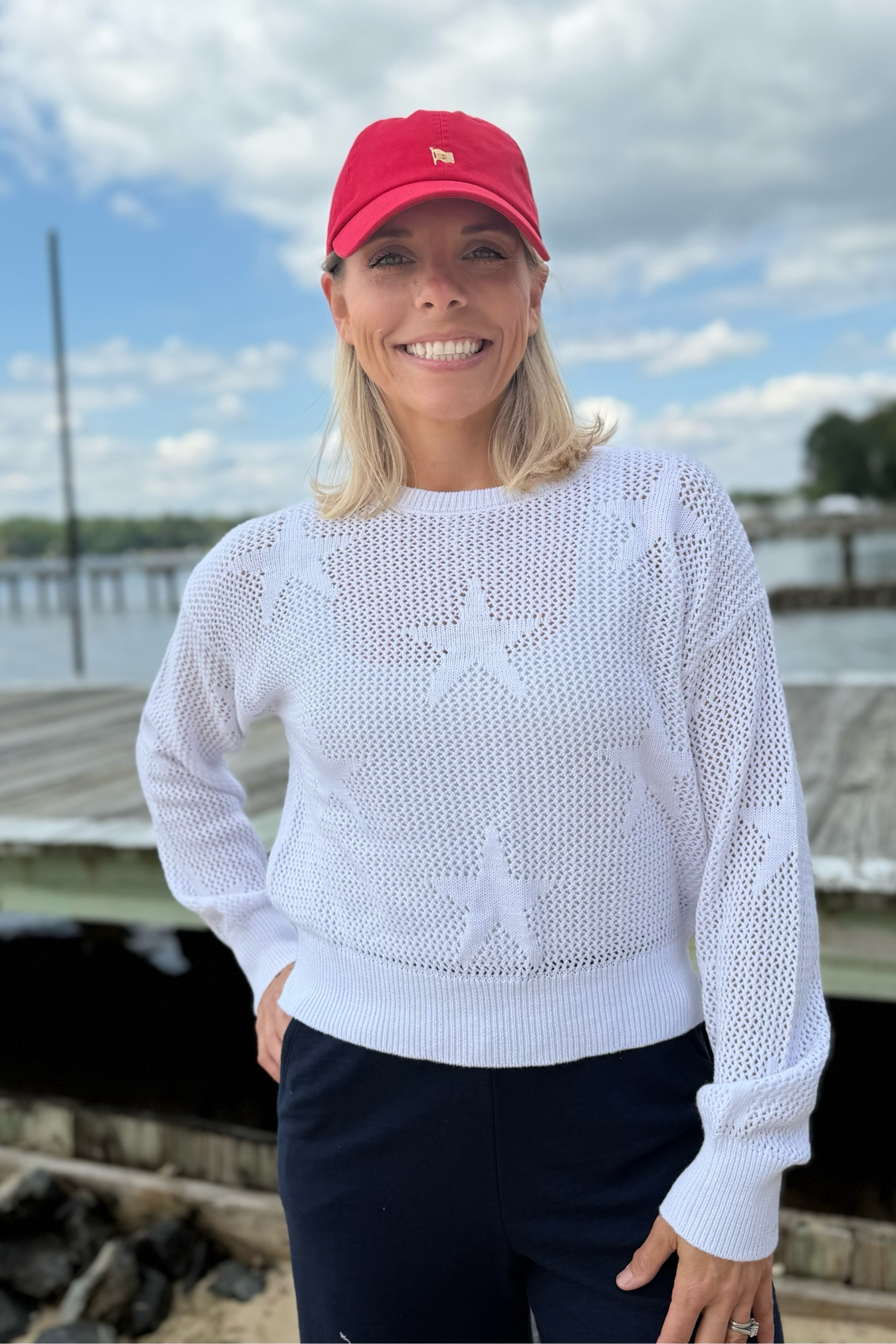 Woman wearing a white sweater with star patterns, a red cap, and navy pants by a dock.