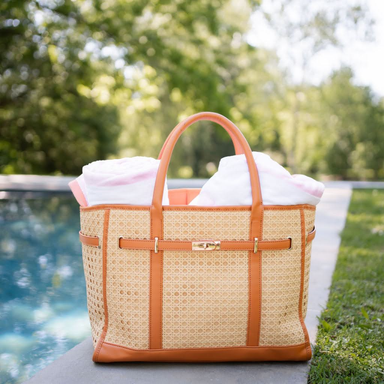 Woven bag with brown handles and trim filled with white towels by a poolside.