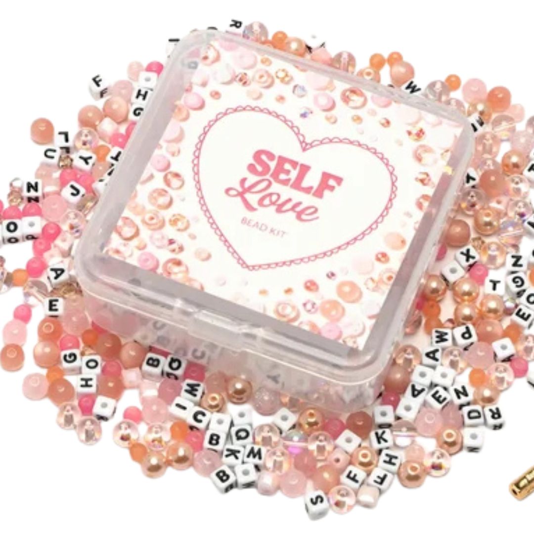 A clear plastic square bead kit container labeled 'SELF Love BEAD KIT' sits on top of a scattered assortment of pink, peach, white, and letter beads on a white background.