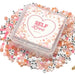 A clear plastic square bead kit container labeled 'SELF Love BEAD KIT' sits on top of a scattered assortment of pink, peach, white, and letter beads on a white background.