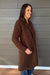 A woman wearing a brown textured bouclé coat stands in front of a gray wood-paneled wall, smiling and looking to the side.