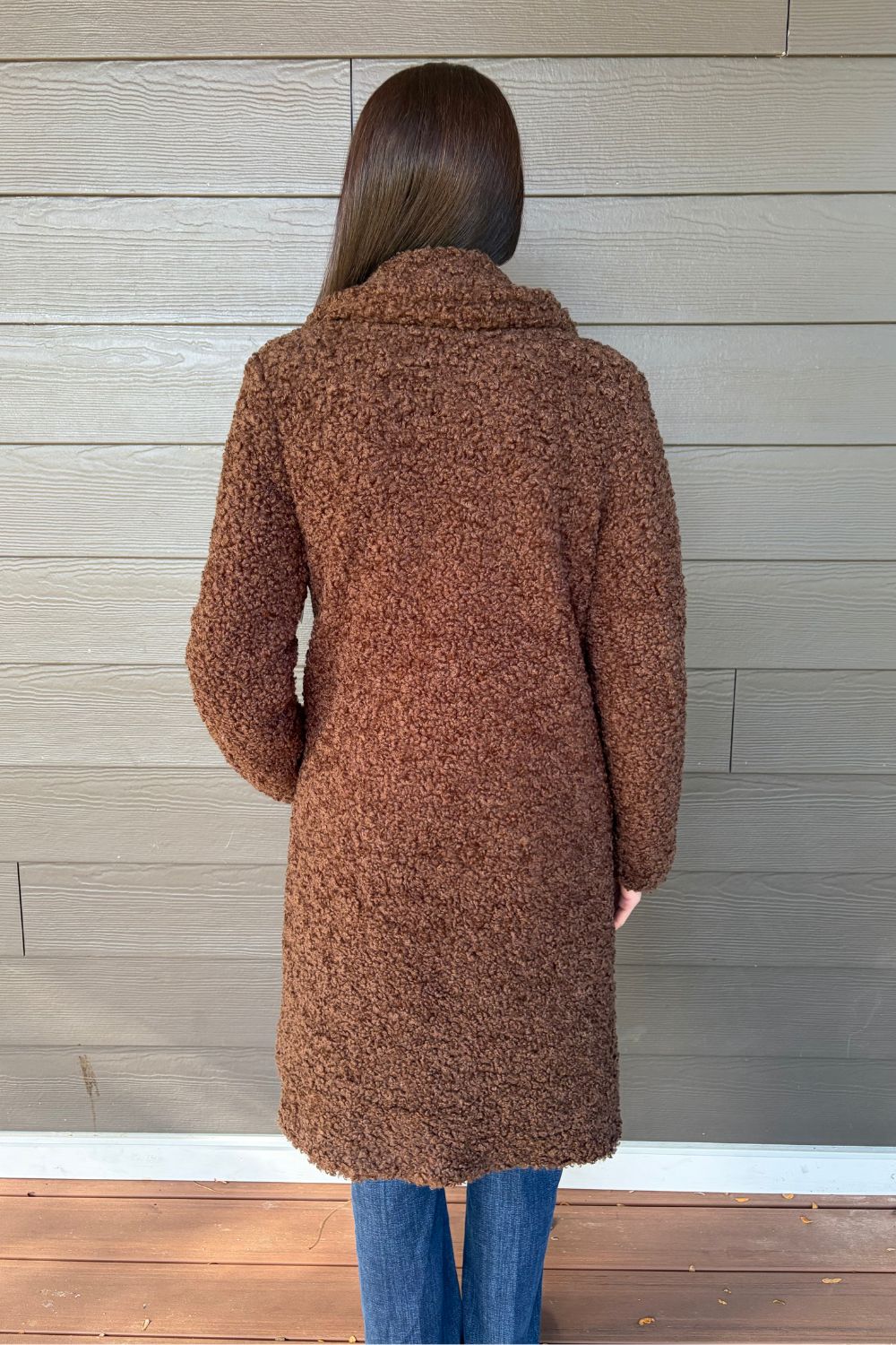 A woman viewed from behind wearing a brown bouclé mid-length coat over blue jeans, standing against a gray wood-paneled wall.