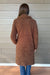 A woman viewed from behind wearing a brown bouclé mid-length coat over blue jeans, standing against a gray wood-paneled wall.