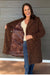 A woman stands on a wooden deck wearing a brown teddy-textured coat and opening it to show a leopard-print lining. She is wearing a cream top and blue jeans against a gray wood-paneled wall.
