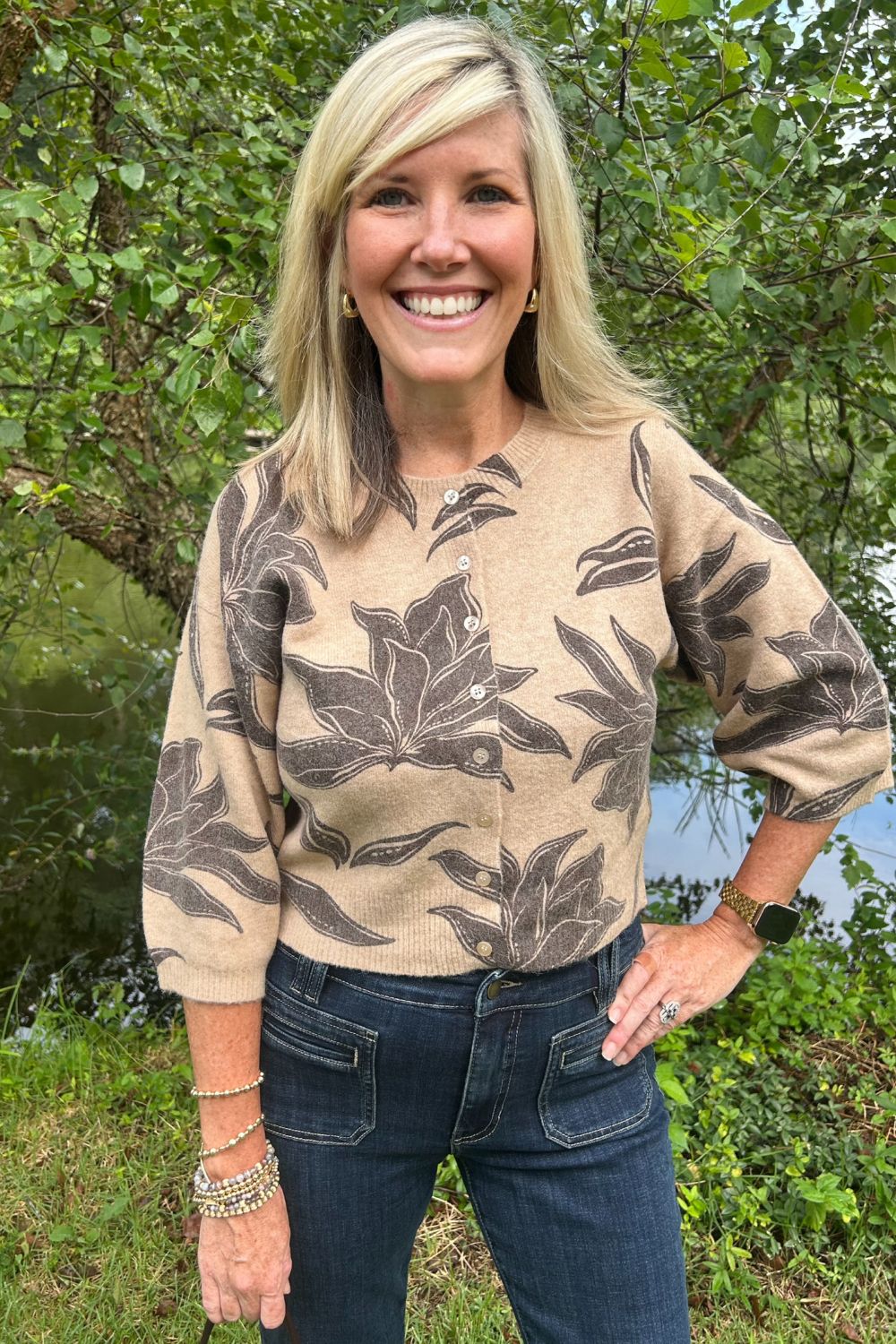 A smiling blonde woman wearing a beige knit long-sleeve top with a dark brown botanical leaf pattern, paired with dark blue jeans, standing outdoors in front of green trees.
