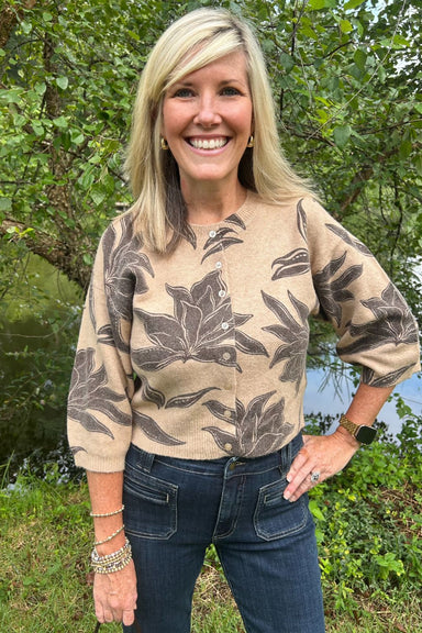 A smiling blonde woman wearing a beige knit long-sleeve top with a dark brown botanical leaf pattern, paired with dark blue jeans, standing outdoors in front of green trees.