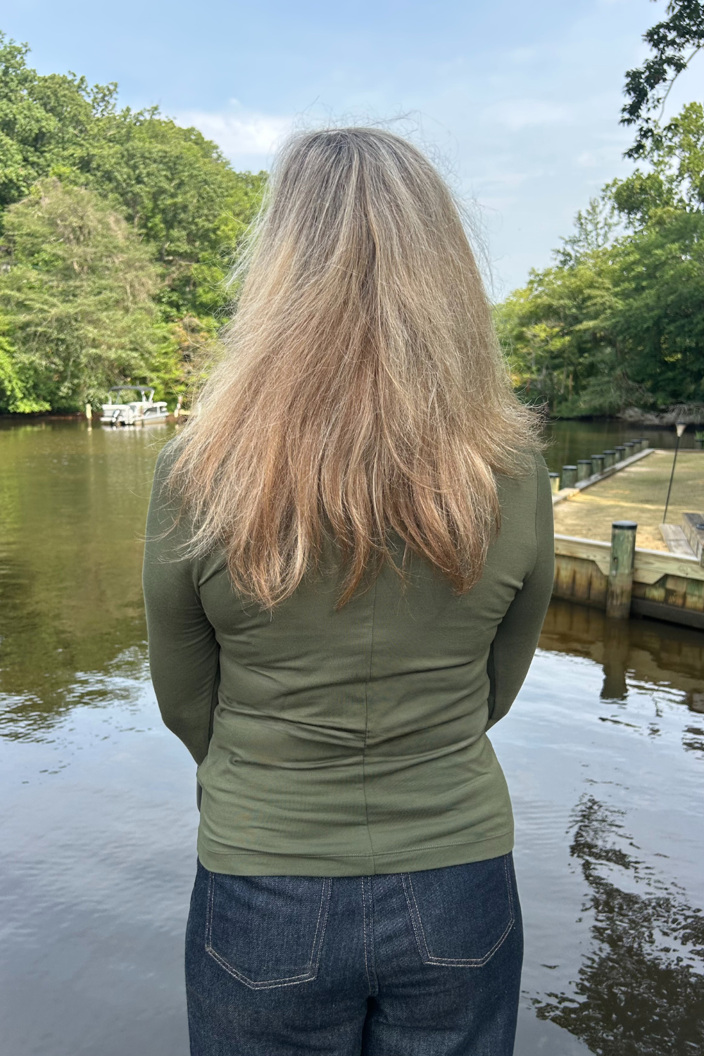 A woman viewed from behind wearing an olive green long-sleeve top and blue jeans, standing outdoors by a body of water with trees and a dock in the background.