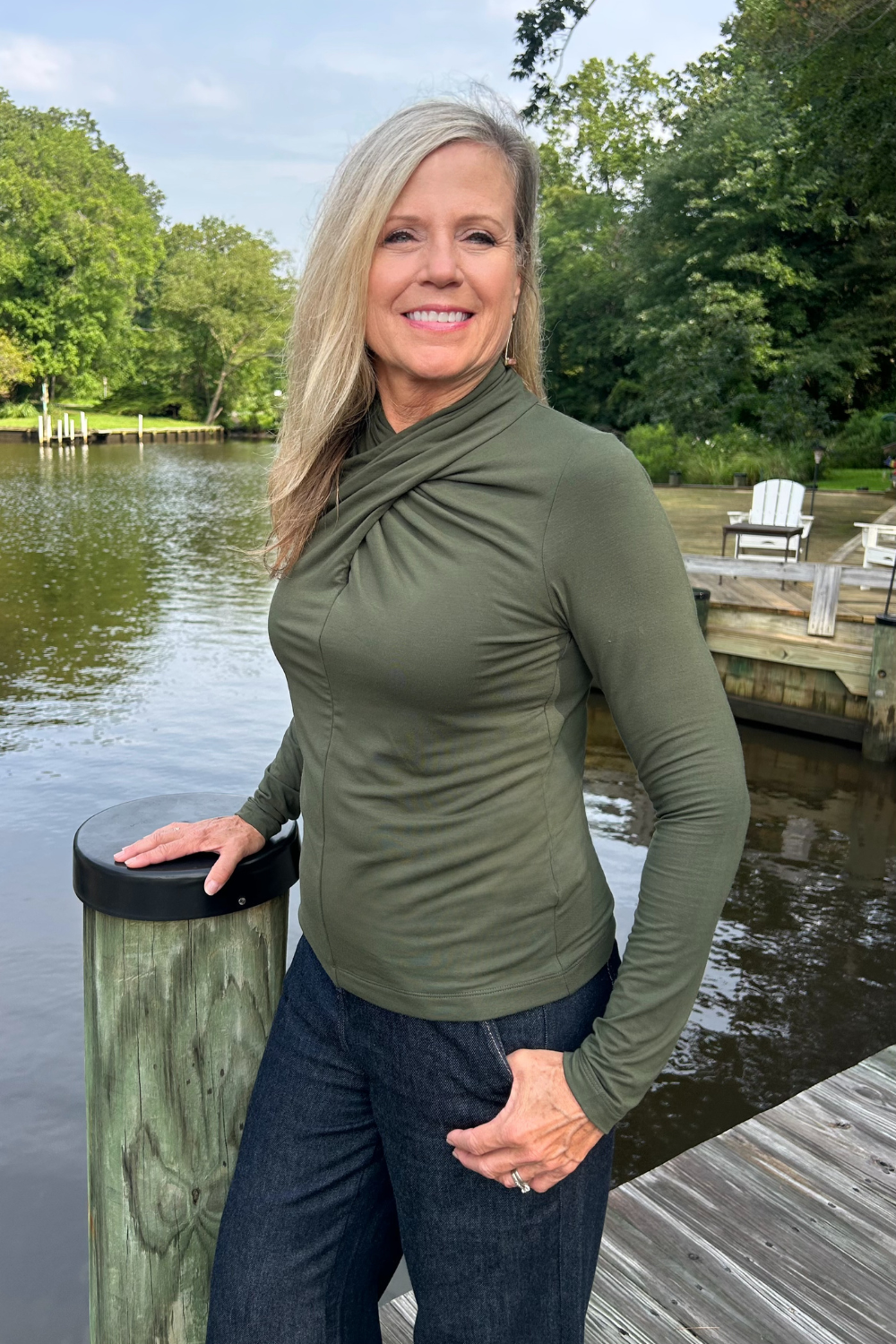 A smiling blonde woman wearing an olive green long-sleeve ruched top and dark jeans stands on a wooden dock next to a lake with trees in the background.