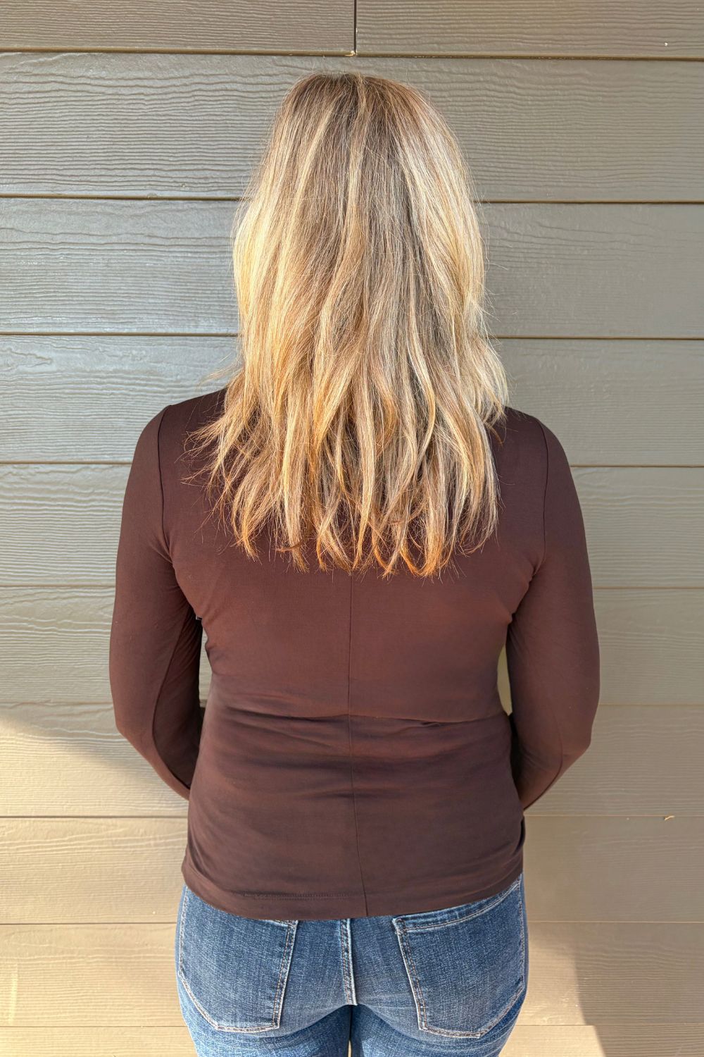 A woman viewed from behind wearing a fitted long-sleeve brown top and blue jeans, standing against a light gray wood-paneled wall.
