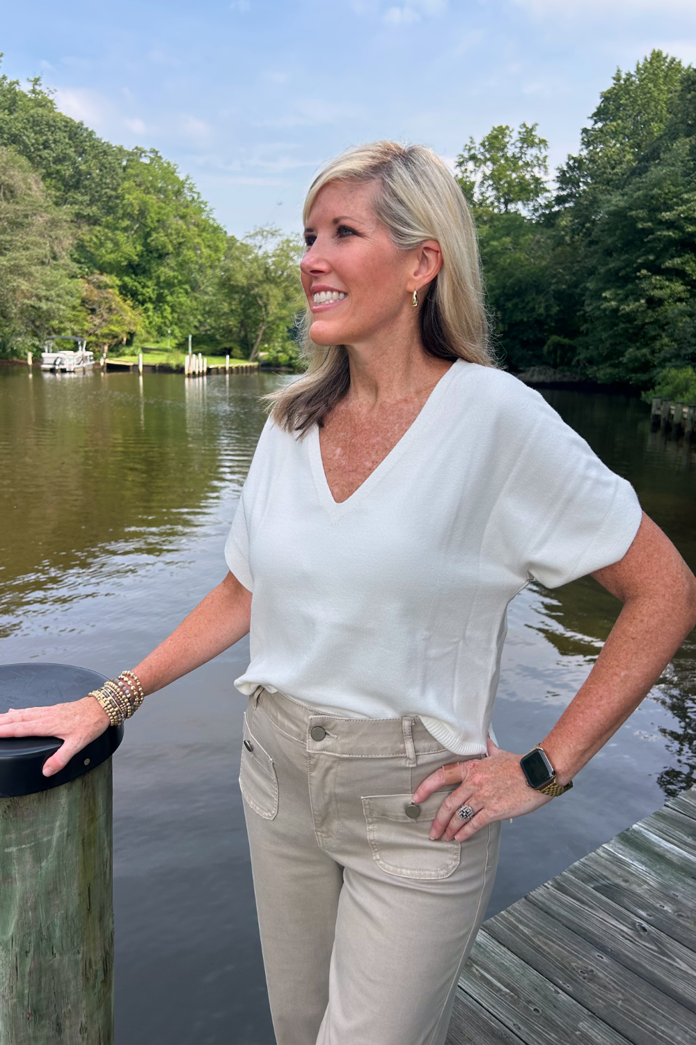 A woman standing on a wooden dock by a calm lake, wearing a white short-sleeve V-neck top and beige pants, with trees and water in the background.
