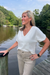 A woman standing on a wooden dock by a calm lake, wearing a white short-sleeve V-neck top and beige pants, with trees and water in the background.